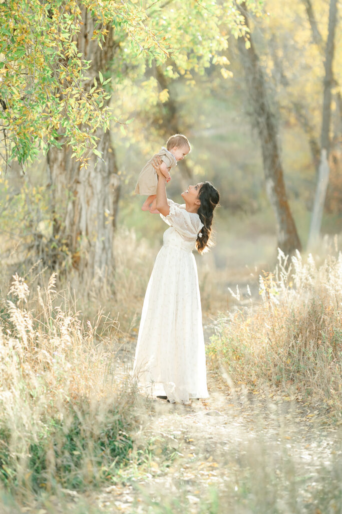 Mother lifting her baby into the air along a tree-lined trail during fall family photos in Snow Basin with warm golden light filtering through the leaves.