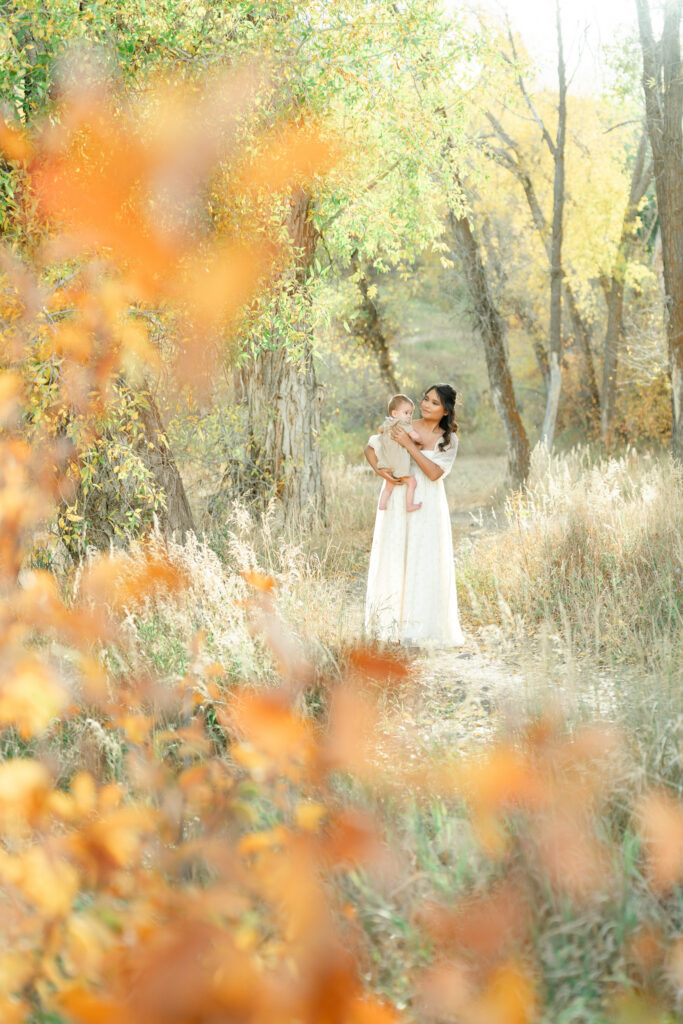 Mother holding her baby in a peaceful autumn forest during Snow Basin fall family photos with vibrant orange leaves framing the foreground.