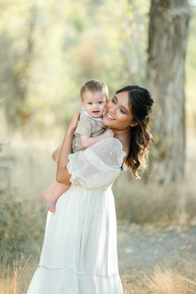 Snow Basin fall family photos of a mother holding her smiling one year-old son in a flowing cream dress beneath glowing autumn trees.