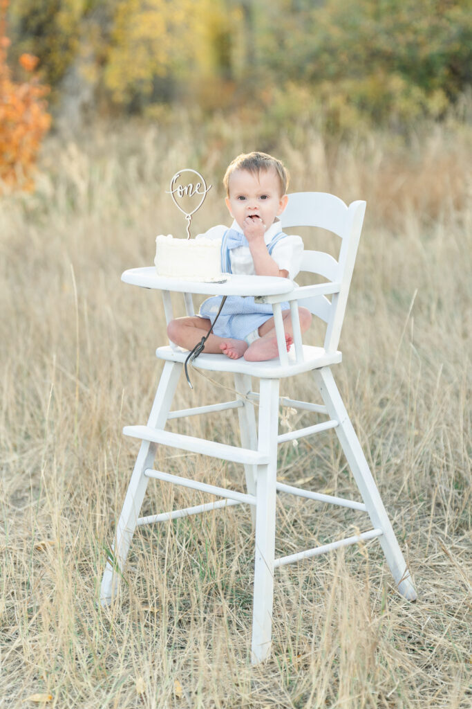 Snow Basin fall family photos of a one-year-old boy enjoying a cake smash while sitting in a white high chair surrounded by golden autumn grass during sunset.