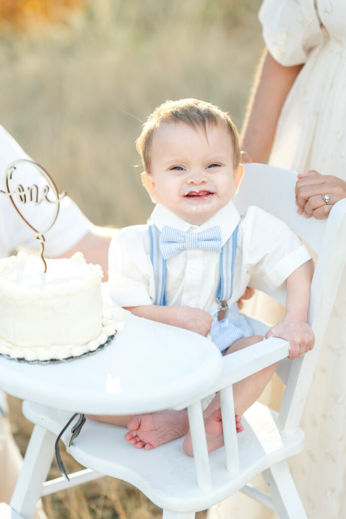 Close-up from a Snow Basin fall family photos session of a smiling one-year-old boy wearing blue suspenders and a bow tie beside his first birthday cake.