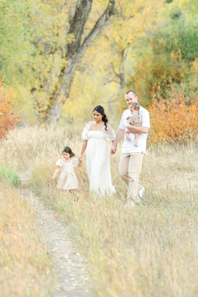Snow Basin fall family photos of a family of four walking hand in hand along a scenic trail surrounded by glowing fall leaves at sunset.