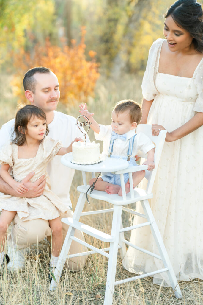 Fall family photos in Snow Basin featuring parents and an older sister celebrating a baby's first birthday together during an outdoor cake smash at golden hour.